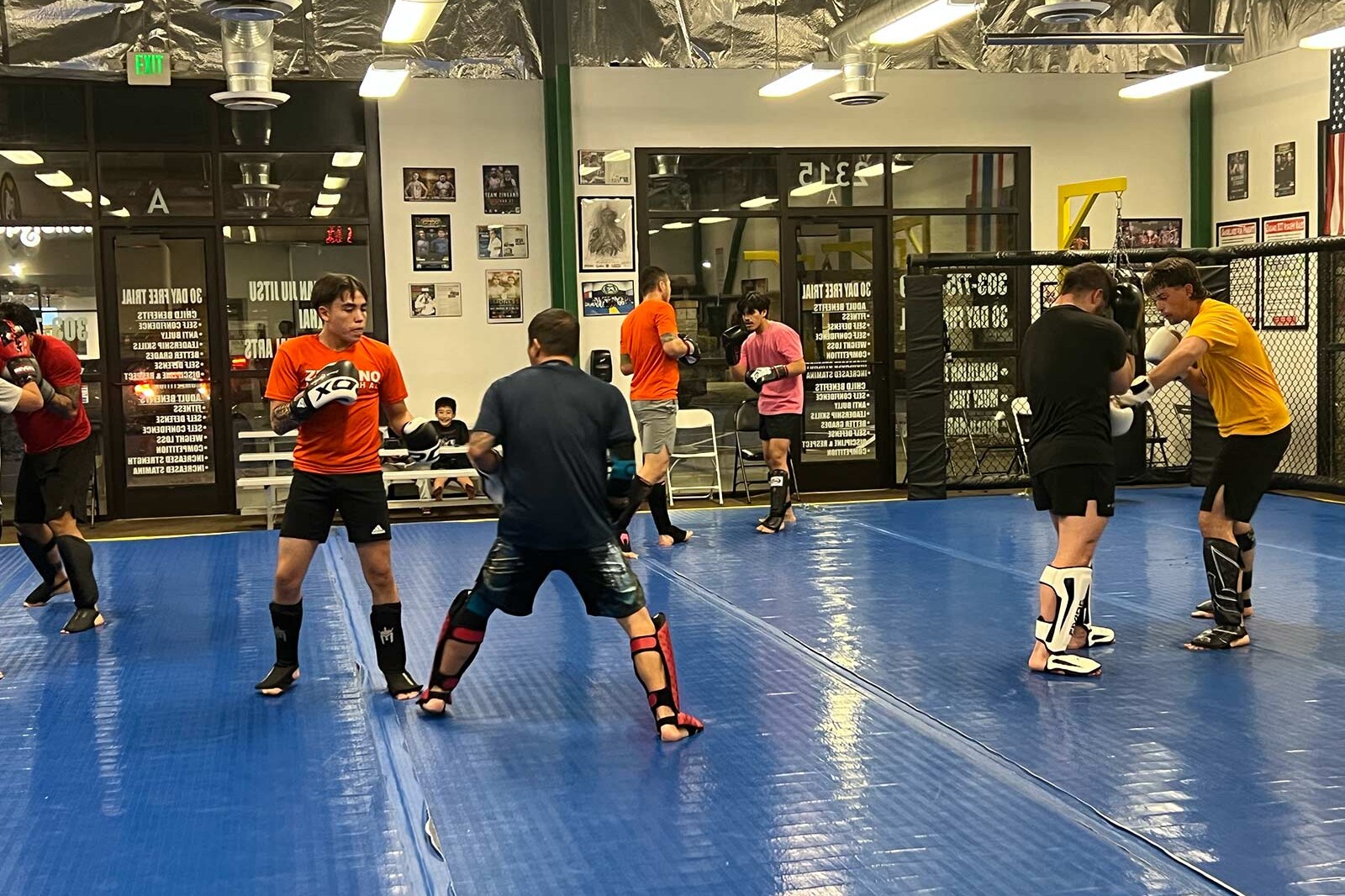 Muay Thai students paired up and practicing striking techniques on blue mats inside a gym, wearing shin guards and gloves, with instructors and spectators watching in the background.