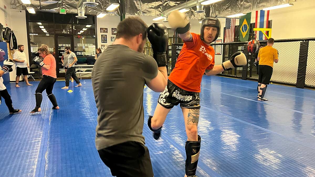Muay Thai students sparring inside a gym, wearing protective gear including gloves, shin guards, and headgear, practicing striking techniques on blue mats with a fenced training area and flags in the background.