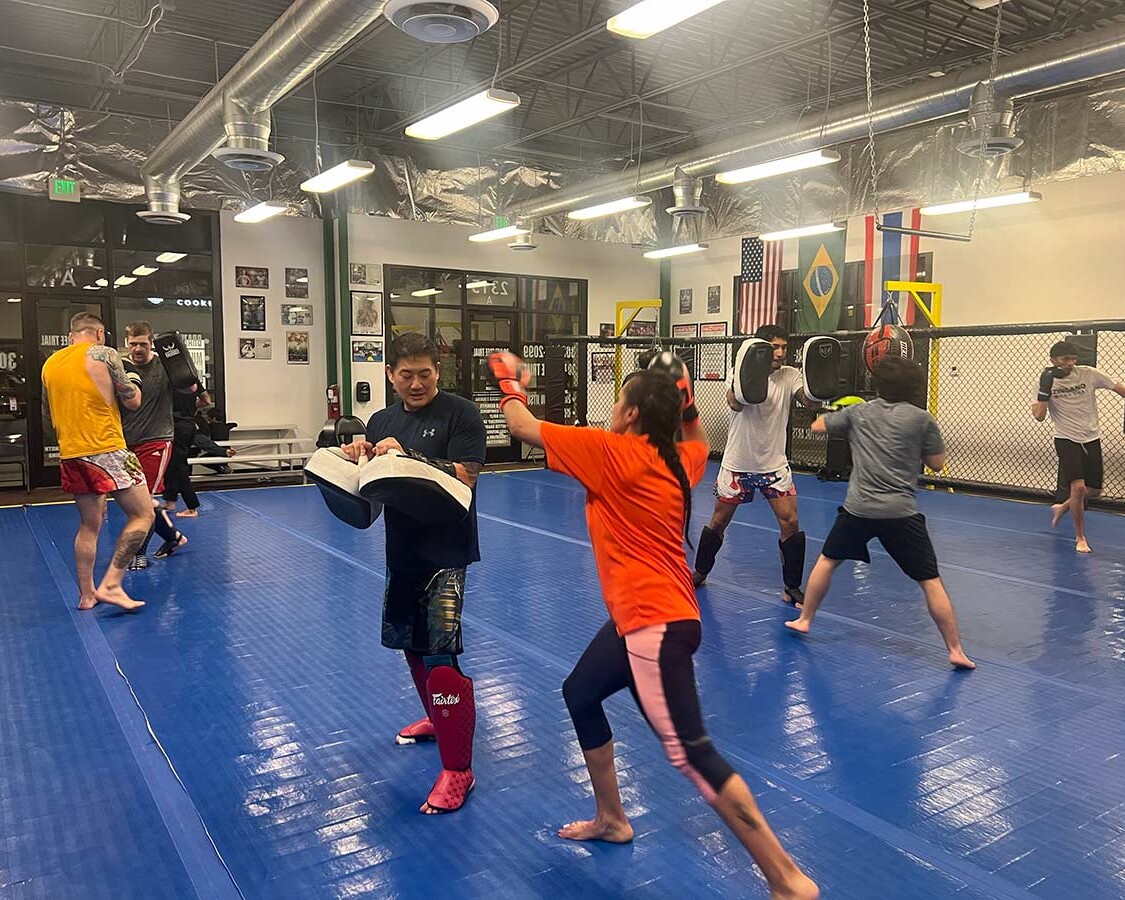 Muay Thai students practicing striking drills with pad holders on blue mats inside Zingano Brighton gym, wearing protective gear, with a fenced training area and international flags in the background.