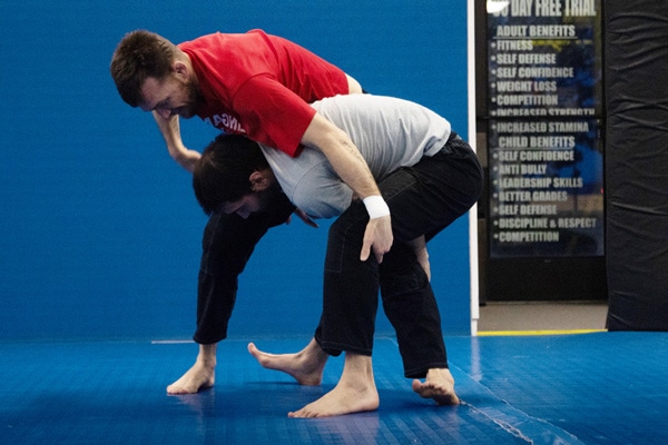 Instructor Cory demonstrating a back control transition during a No-Gi Brazilian Jiu-Jitsu class at Zingano Brighton, using a hands-on technique with a student on the blue mat, with gym benefit signage visible in the background.