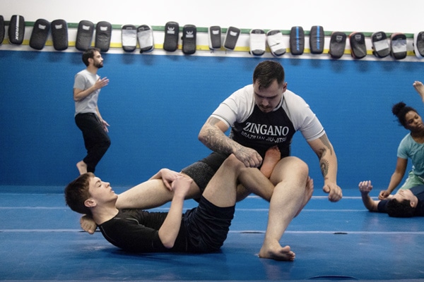 tudents drilling leg lock techniques during a No-Gi Brazilian Jiu-Jitsu class at Zingano Brighton, with a pair practicing positional control on the mat and other participants training in the background under a wall lined with striking pads.